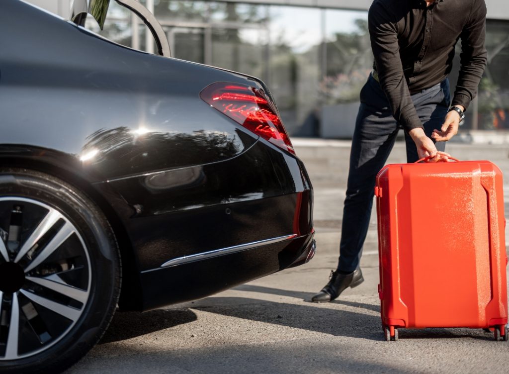 Un chauffeur privé en costume est en train de ranger une valise rouge dans le coffre de sa voiture haut de gamme.
